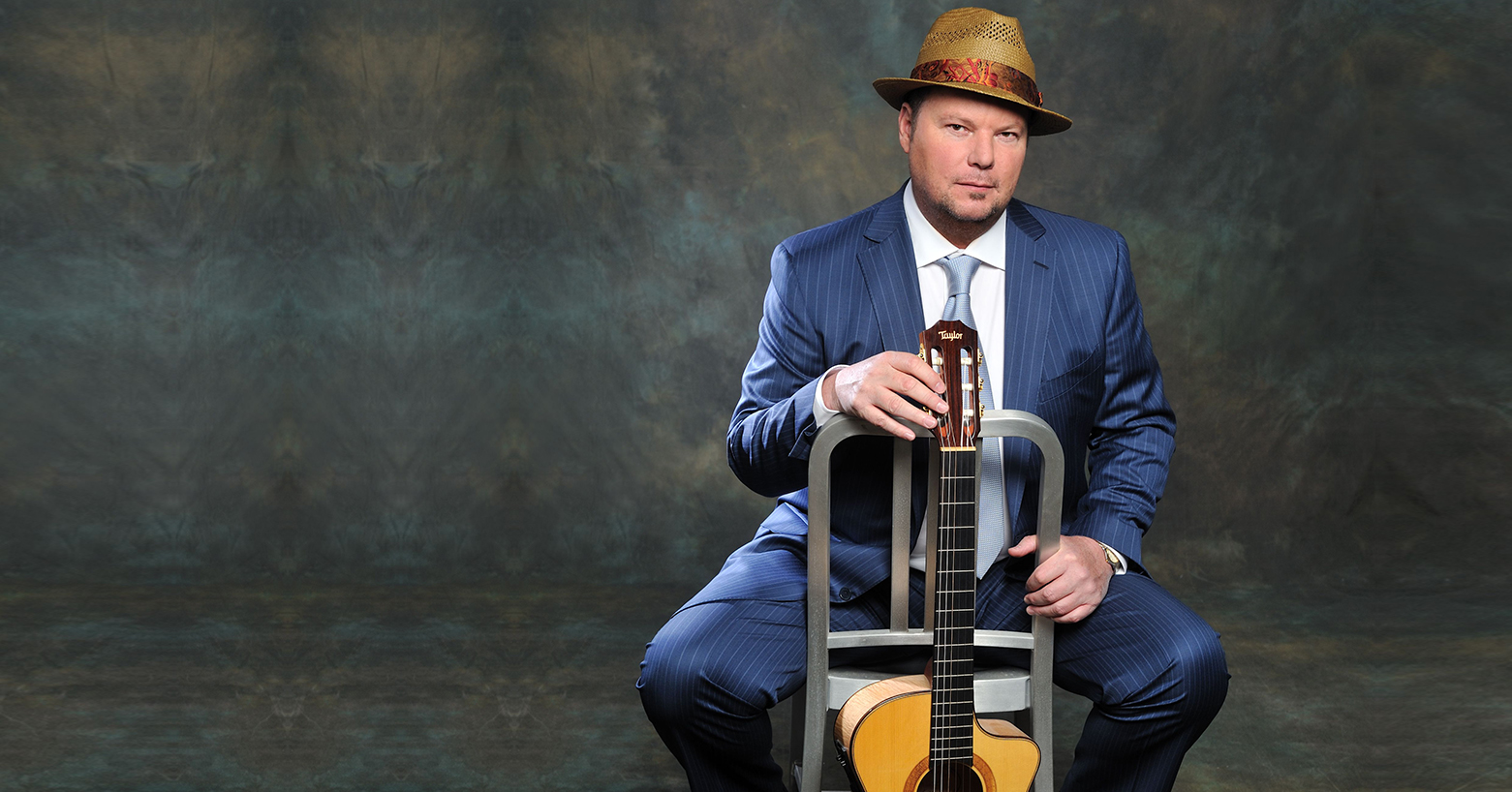 Christopher Cross wearing a blue pinstriped suit with a straw hat holding a guitar.