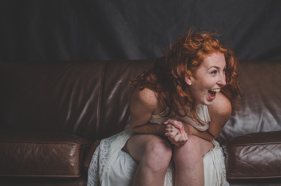 A woman with redhair laughing while wearing a white dress and sitting on a brown leather couch.