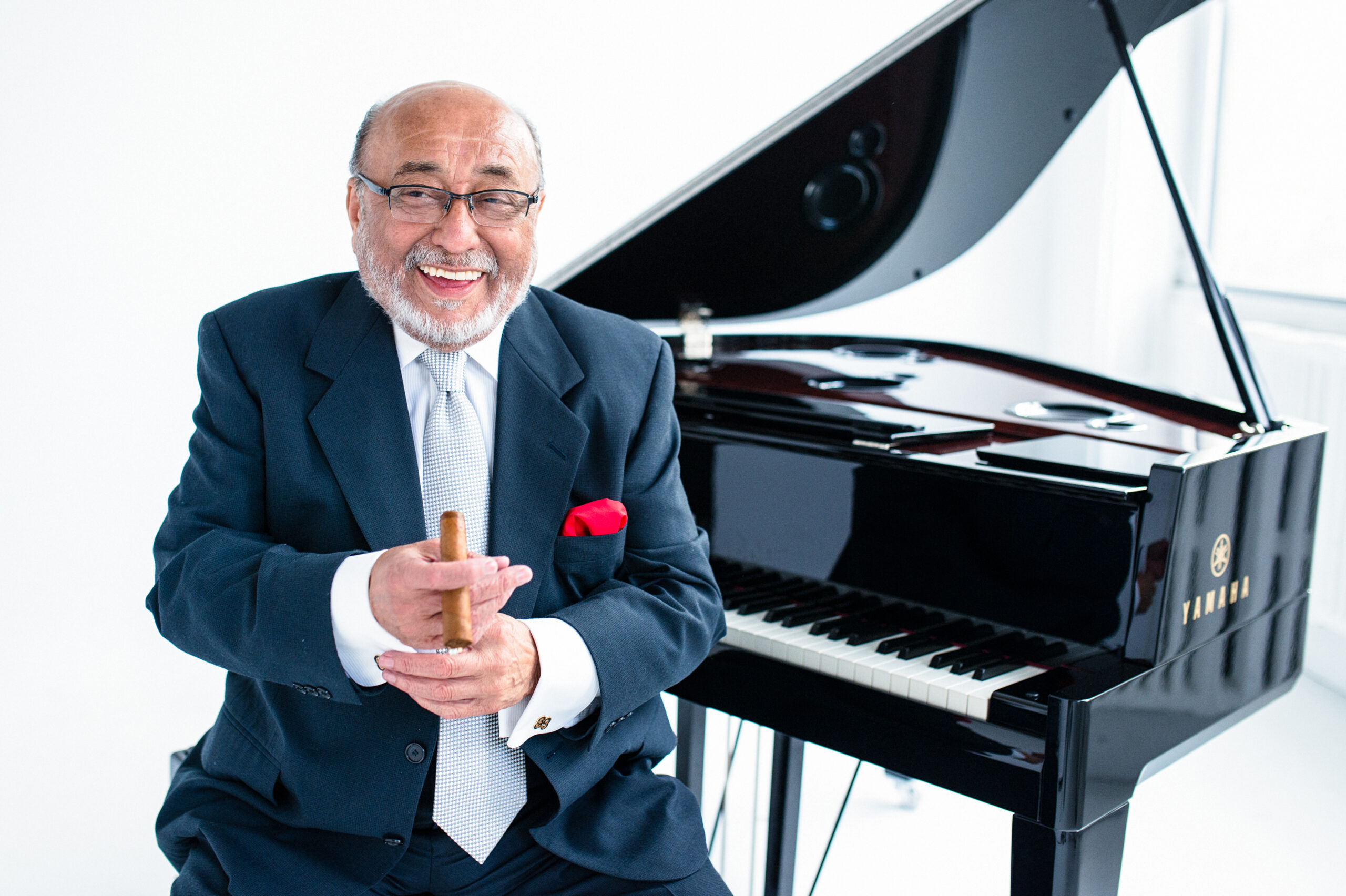 Eddie Palmieri holding a cigar, smiling and sitting in front of a piano.
