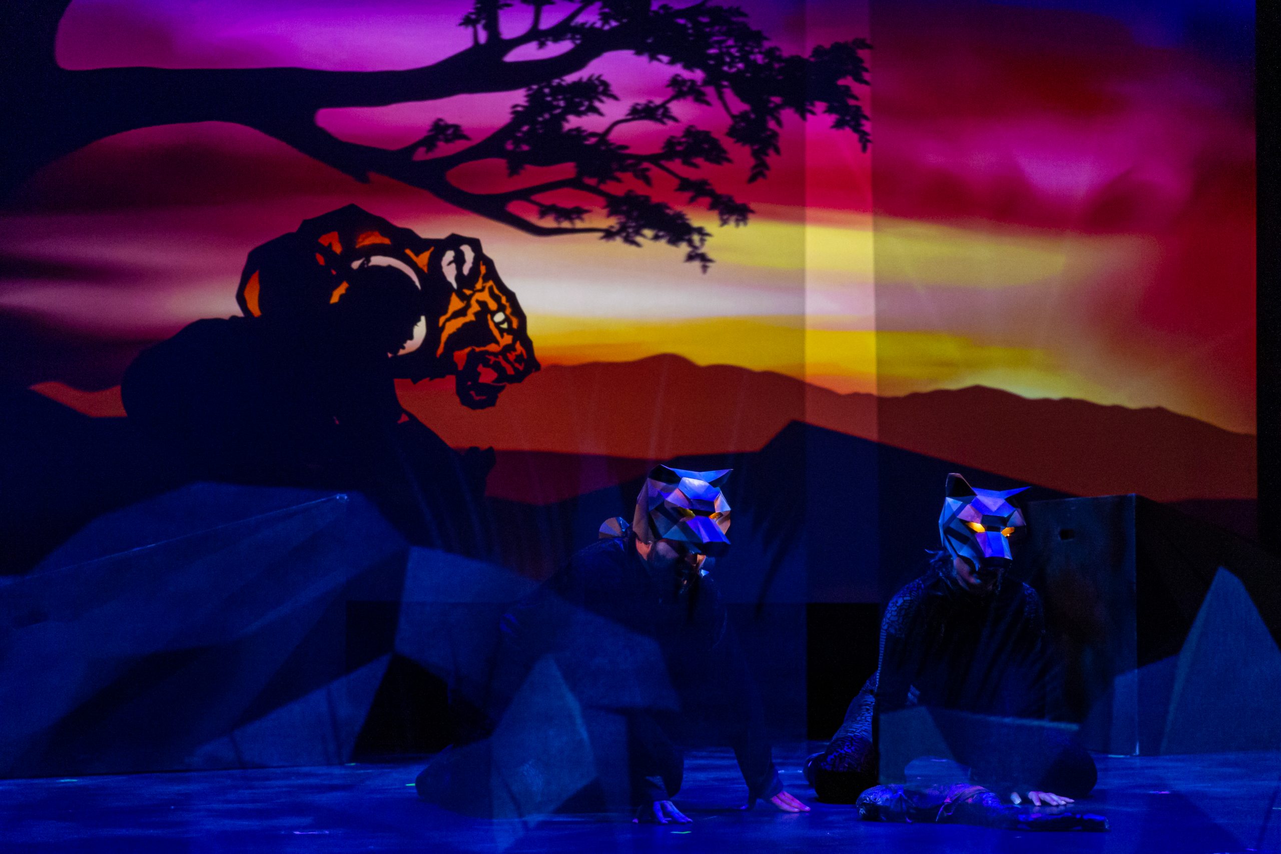 Two cast members wearing wolf masks with glowing eyes. A shadow puppet of Shere Khan approaches from the left.