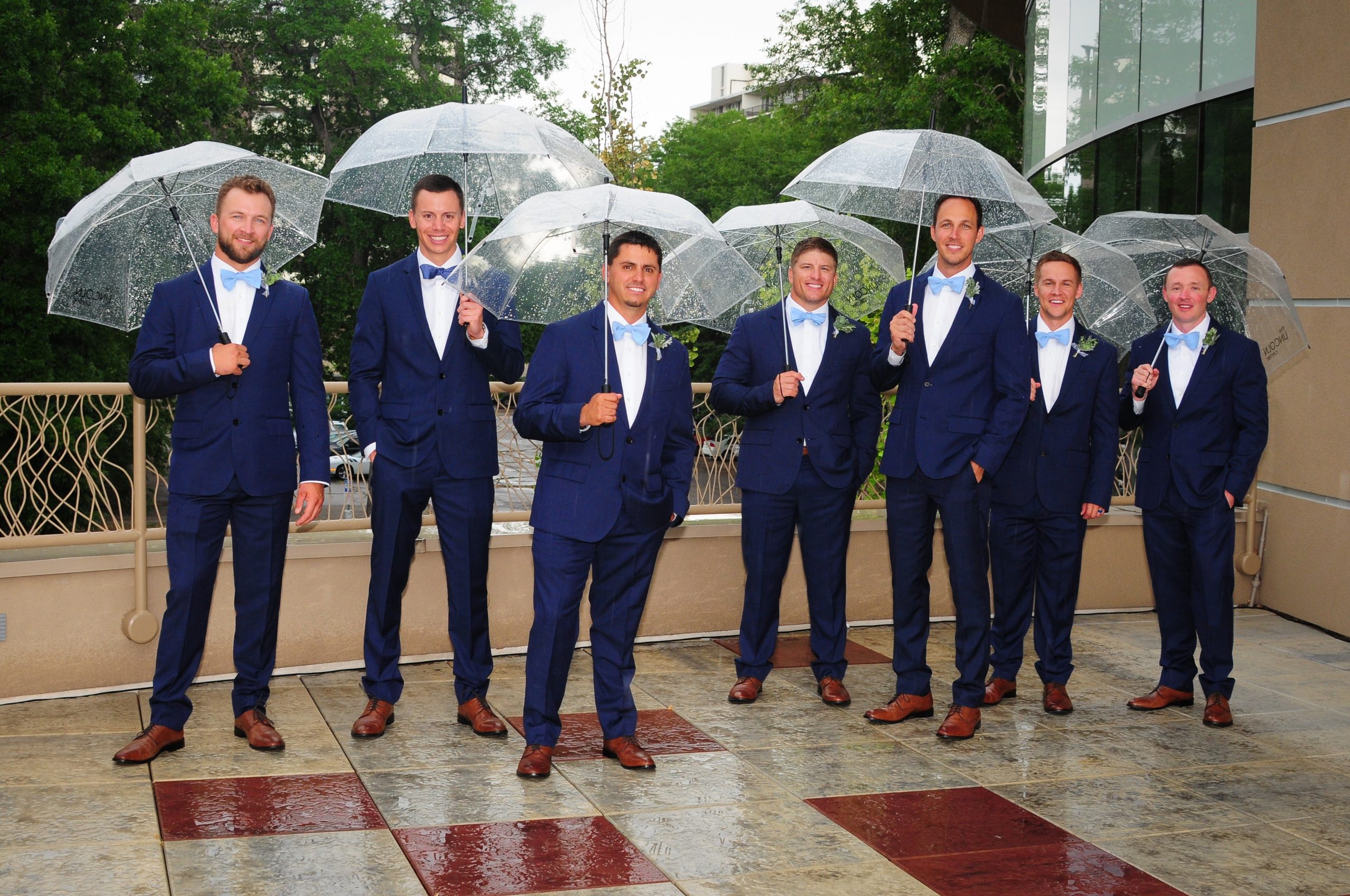 Seven groomsmen in blue suits on the rooftop deck holding clear umbrellas.