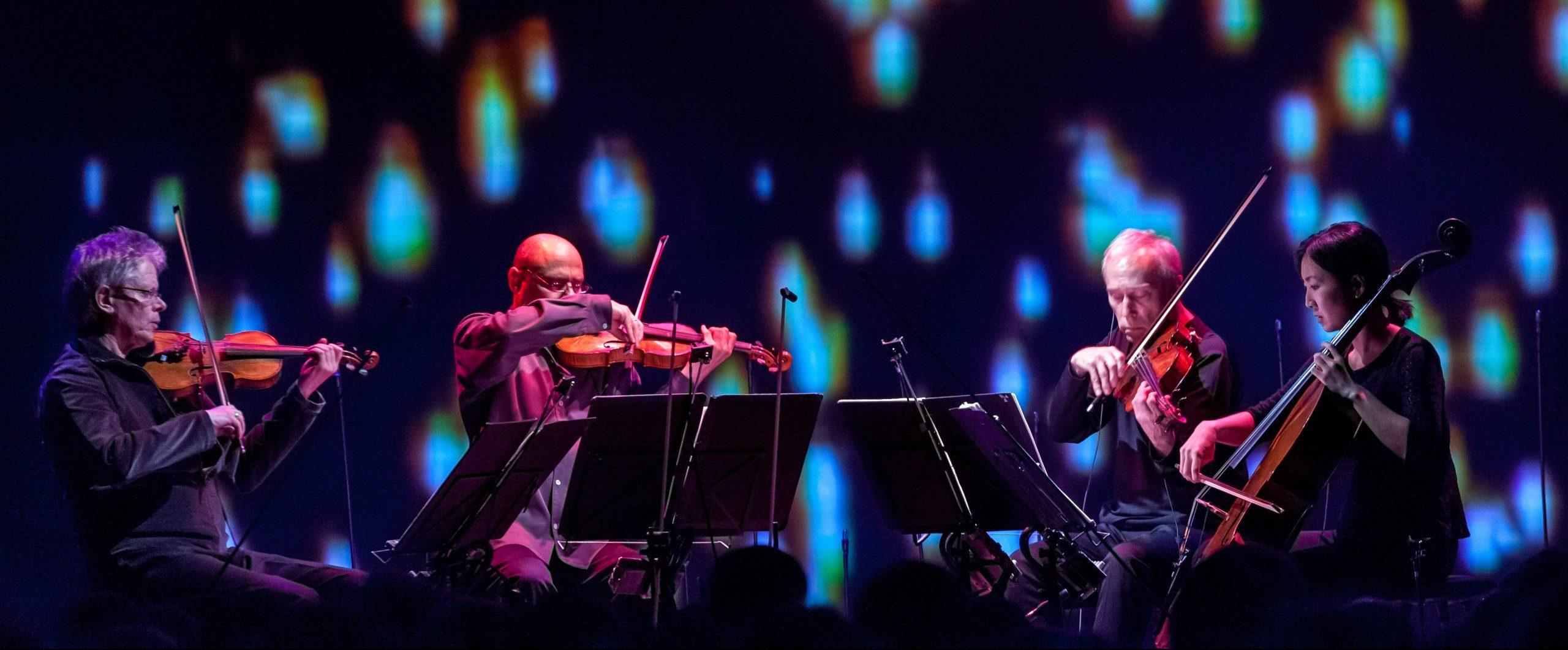 The members of Kronos Quartet playing on stage in front of a background that looks like embers.