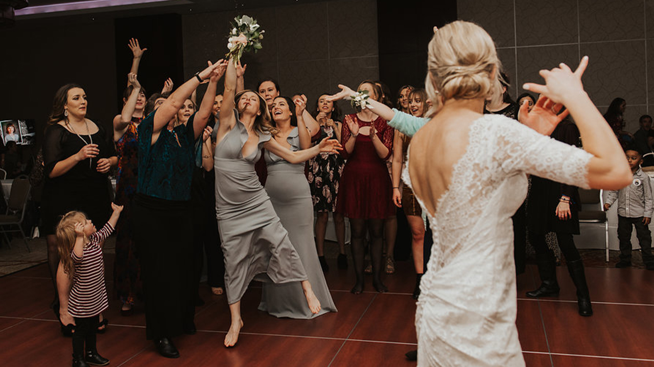 A bride performs a bouquet toss as guests jump to catch it at a wedding reception.