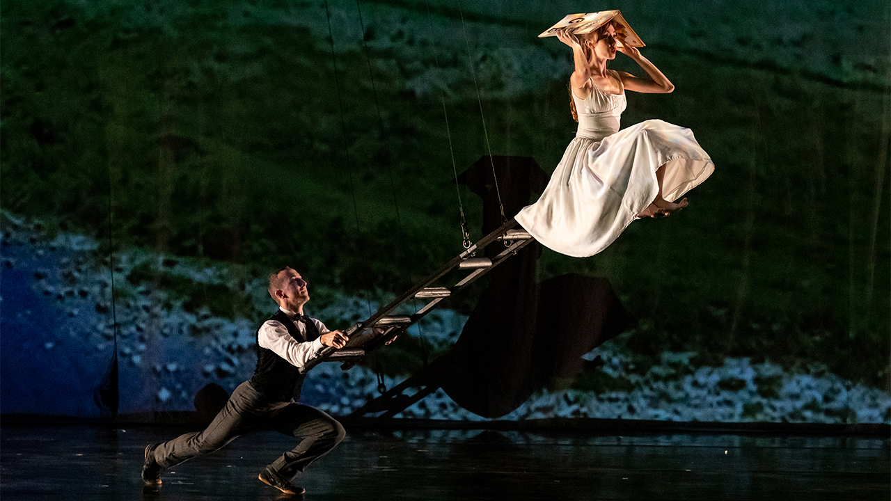 Momix dancers portraying Alice in Wonderland. Alice is reading a book a top of a ladder suspended diagonally in midair.