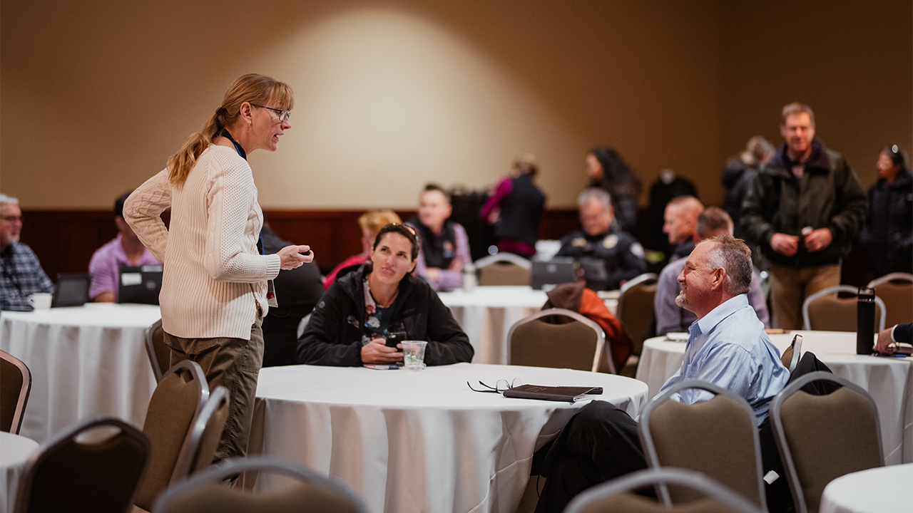 A meeting presenter speaks with attendees at a table in Columbine Room.