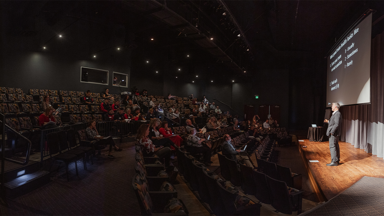 A speaker stands on the stage of Magnolia Theatre and presents to attendees in front of a projection screen.