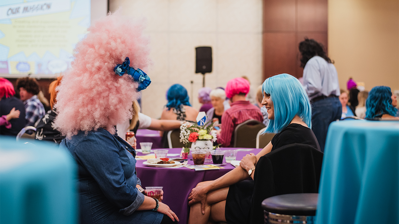 Women in colorful wigs sit and talk at a table.