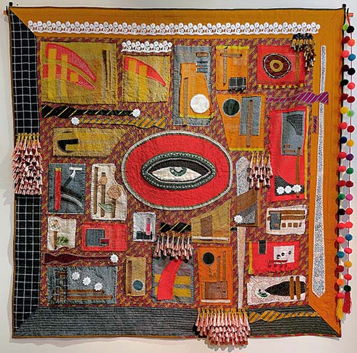 quilt with a large eye in the center and mixed media elements such as beads and metal cones attached