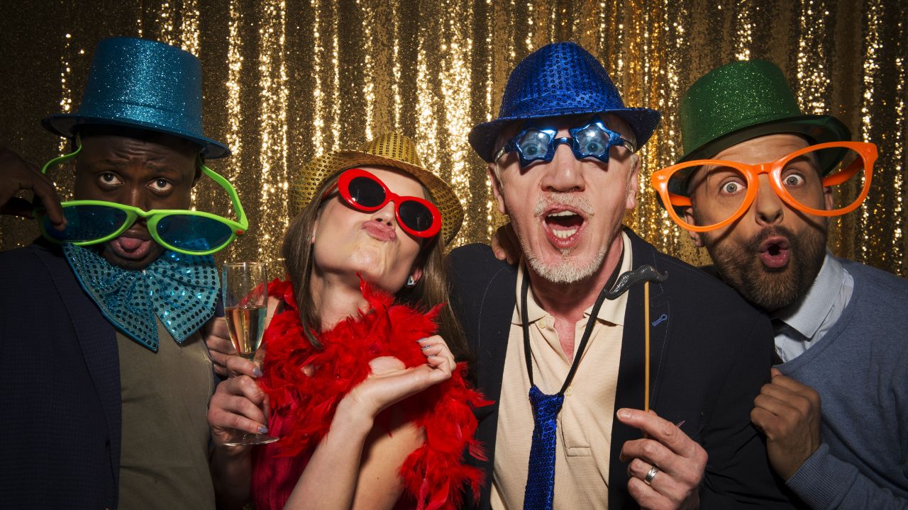 Four people laughing and wearing funny sunglasses and hats in front of a gold background.
