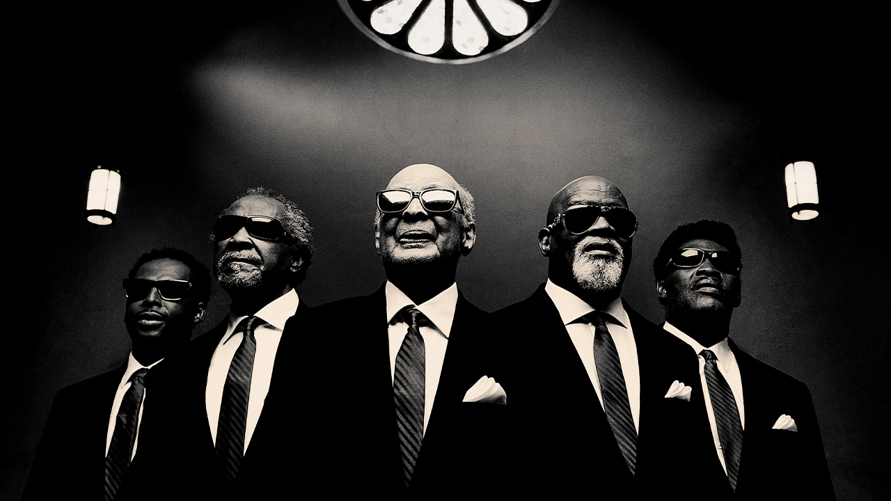 A black and white photo of the Blind Boys standing beneath stained glass.