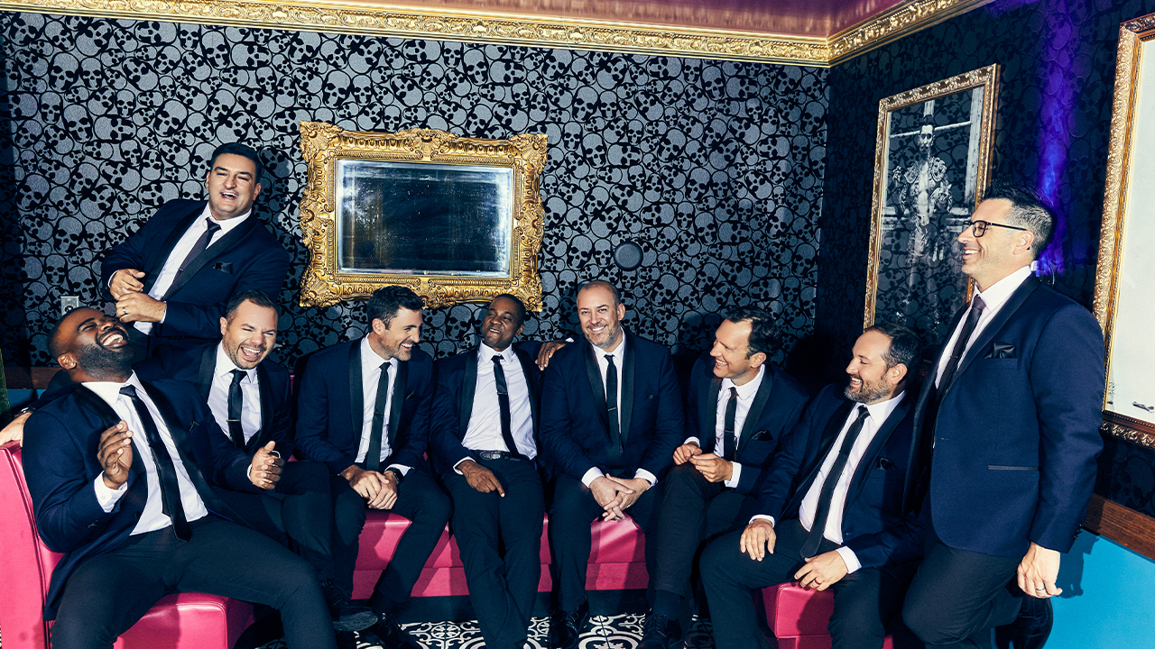 Photo of the members of Straight No Chaser wearing suits and siting in a lounge with skull wallpaper on a pink couch.