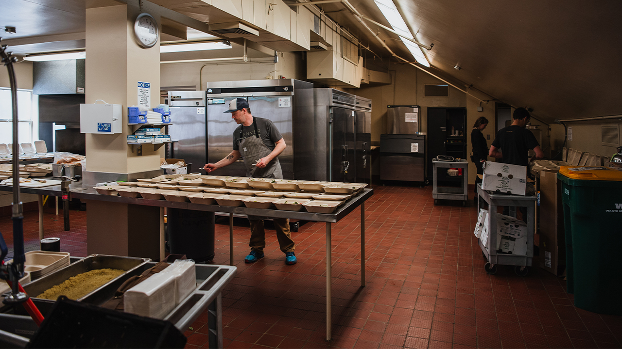 People work in a commercial kitchen with multiple refrigerators, sinks and prep counters.
