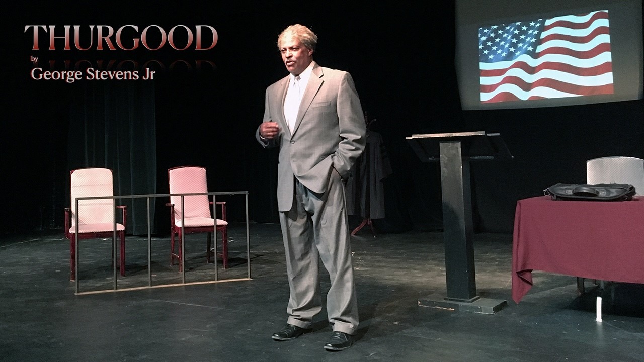 A grey haired man in a grey suit stands on stage flanked by a jury box and an American flag. Text in upper left corner says: George Stevens Jr.