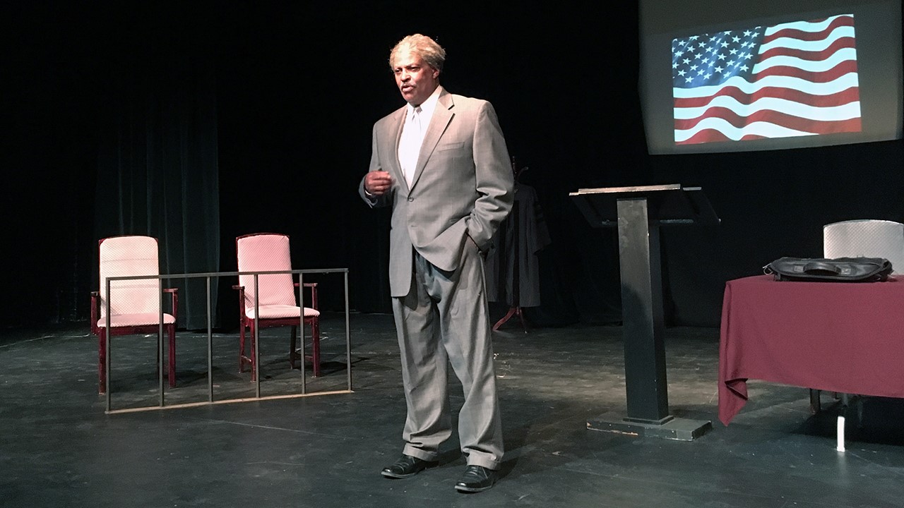 A grey haired man in a grey suit stands on stage near a jury box and an American flag.