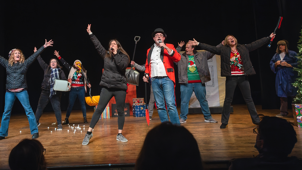 Actors singing and dancing on stage in holiday sweaters while holding snow removal equipment.