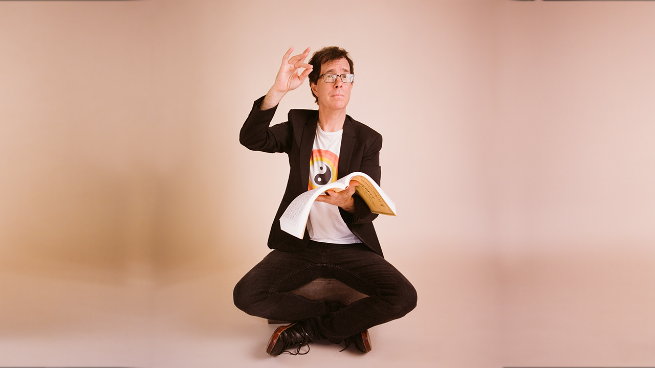 Ben Folds in front of a light coral background sitting cross-legged and holding a book of sheet music.