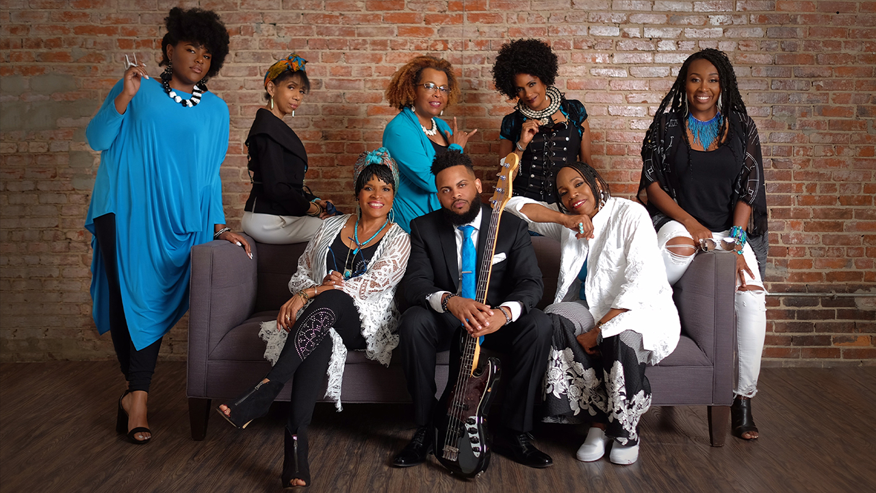 Eight members of Sweet Honey in the Rock sitting in front of a brick wall.