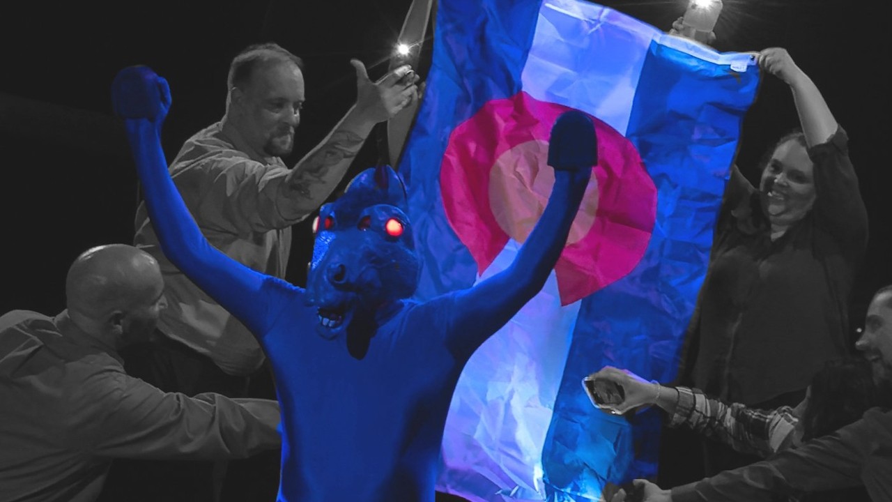A man in a Bluecifier Blue Horse costume stands in front of a Colorado flag with his arms raised in celebration.