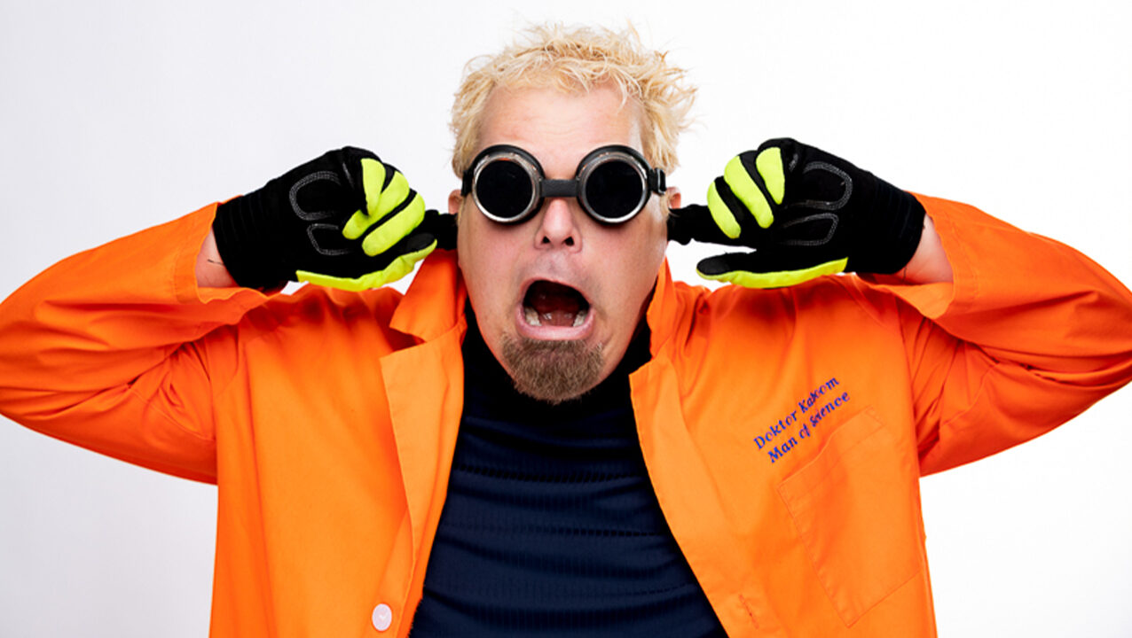 Doktor Kaboom wears a bright orange jacket and has his fingers in his ears in preparation for an explosion