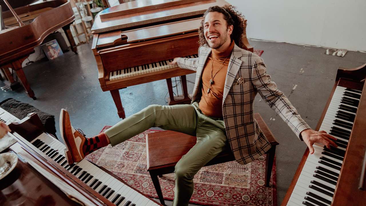 Emmet Cohen in a plaid suit smiling and stretched out playing 3 pianos