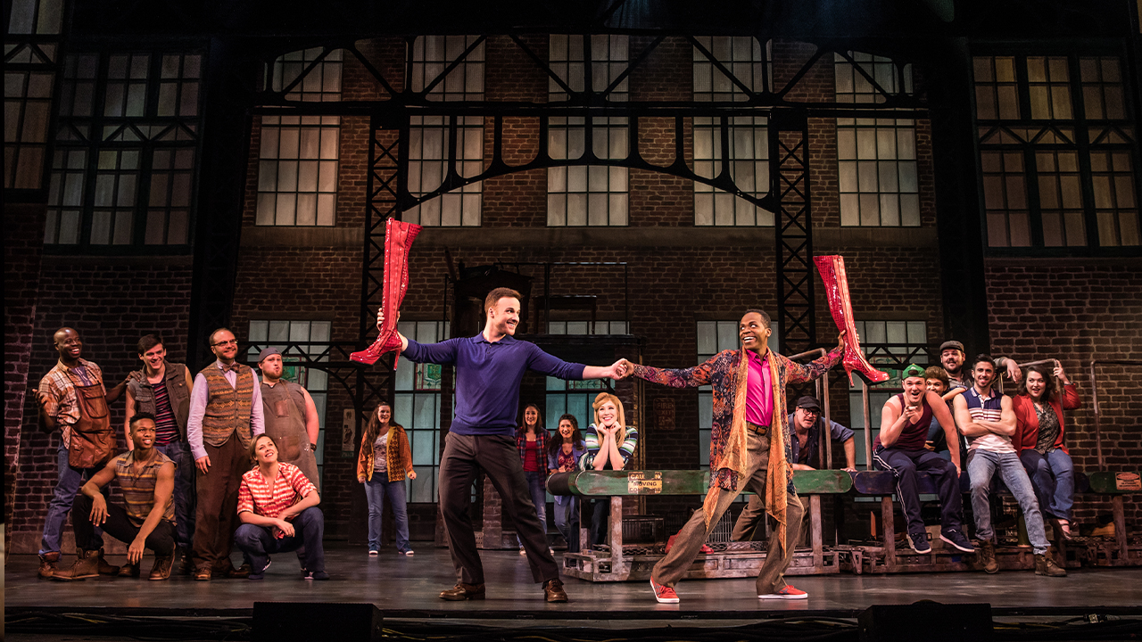 The two main characters of Kinky Boots stand on stage hand in hand with their big red high heel pumps