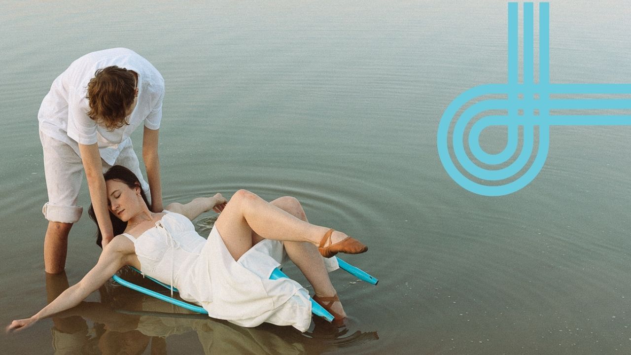 A male presenting dancer holds a chair with a female presenting dancer in a lake as if they are performing a baptism.