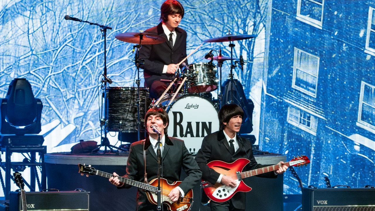 Members of RAIN (The Beatles tribute band) stand on stage playing their instruments with the classic early Beatles black suits and bowl cuts