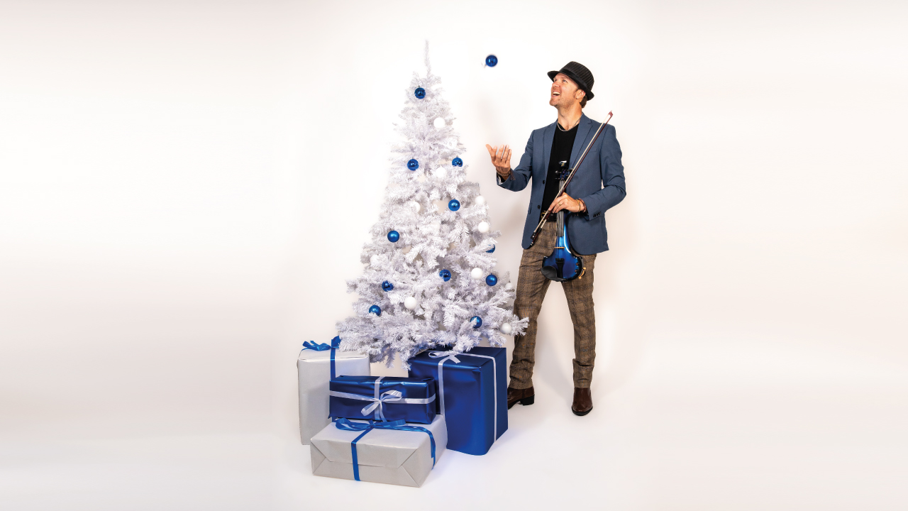 Man holding a blue violin stands next to a white Christmas tree with blue decorations and blue presents under the tree.