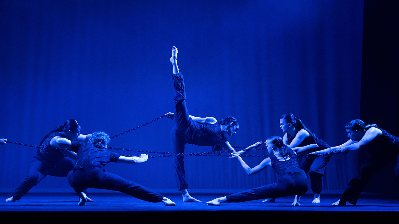 Dancers on stage performing dramatic choreography in deep blue lighting.