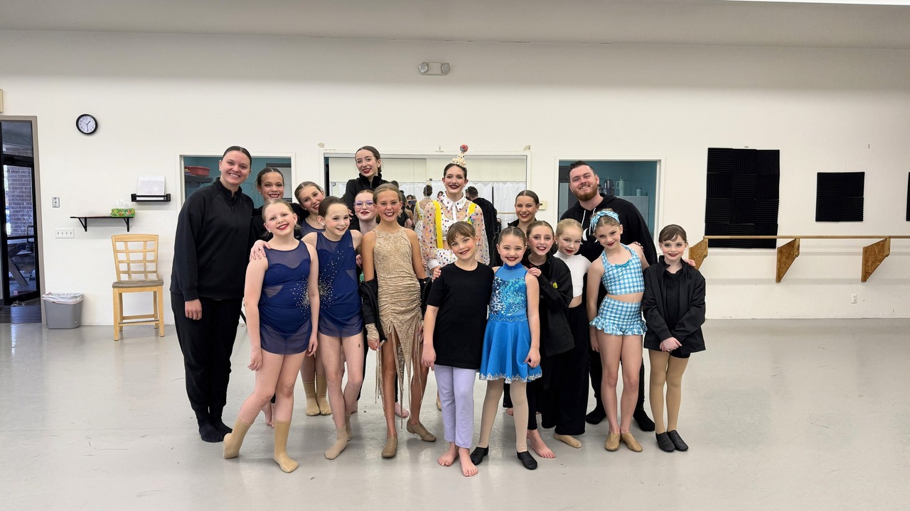 A group of student dancers in various costumes and team apparel pose together with two instructors in a dance studio during a dress rehearsal.