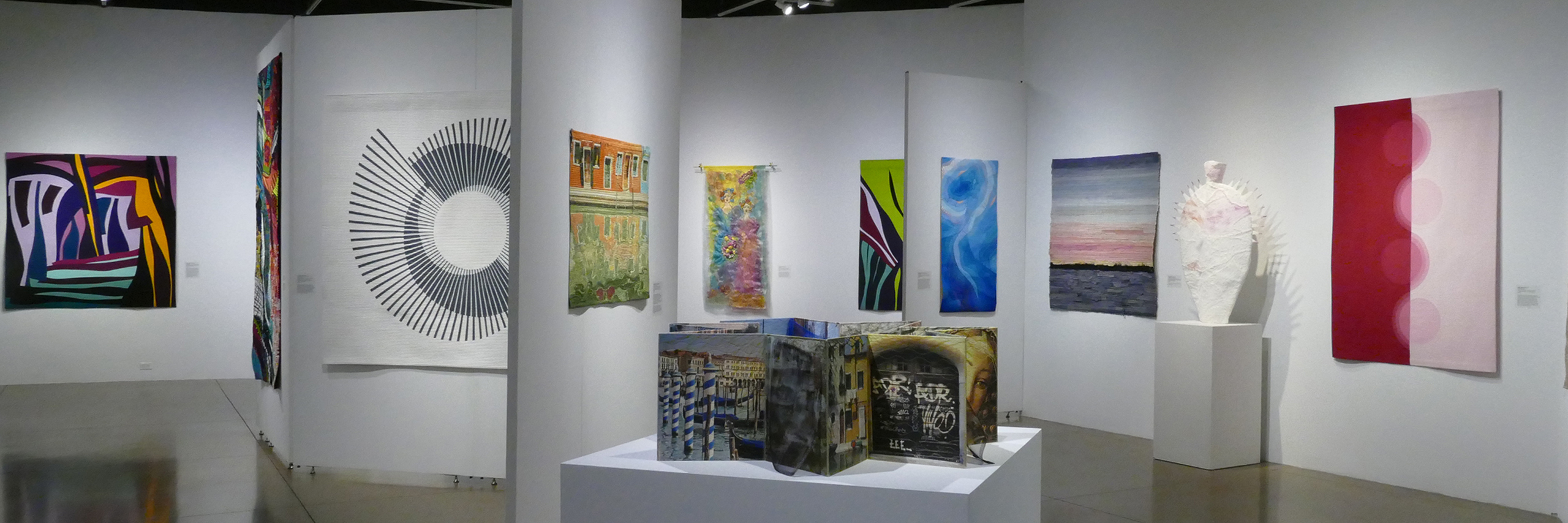 Image shows a gallery with a variety of multi colored artistic quilts hung on walls that are placed in at different angles in the space