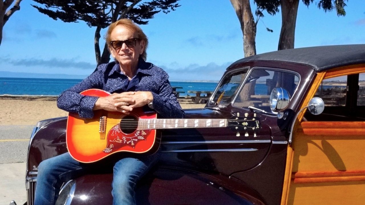 Picture of Al Jardine sitting on an 1970s "woodie" car on a beach in California