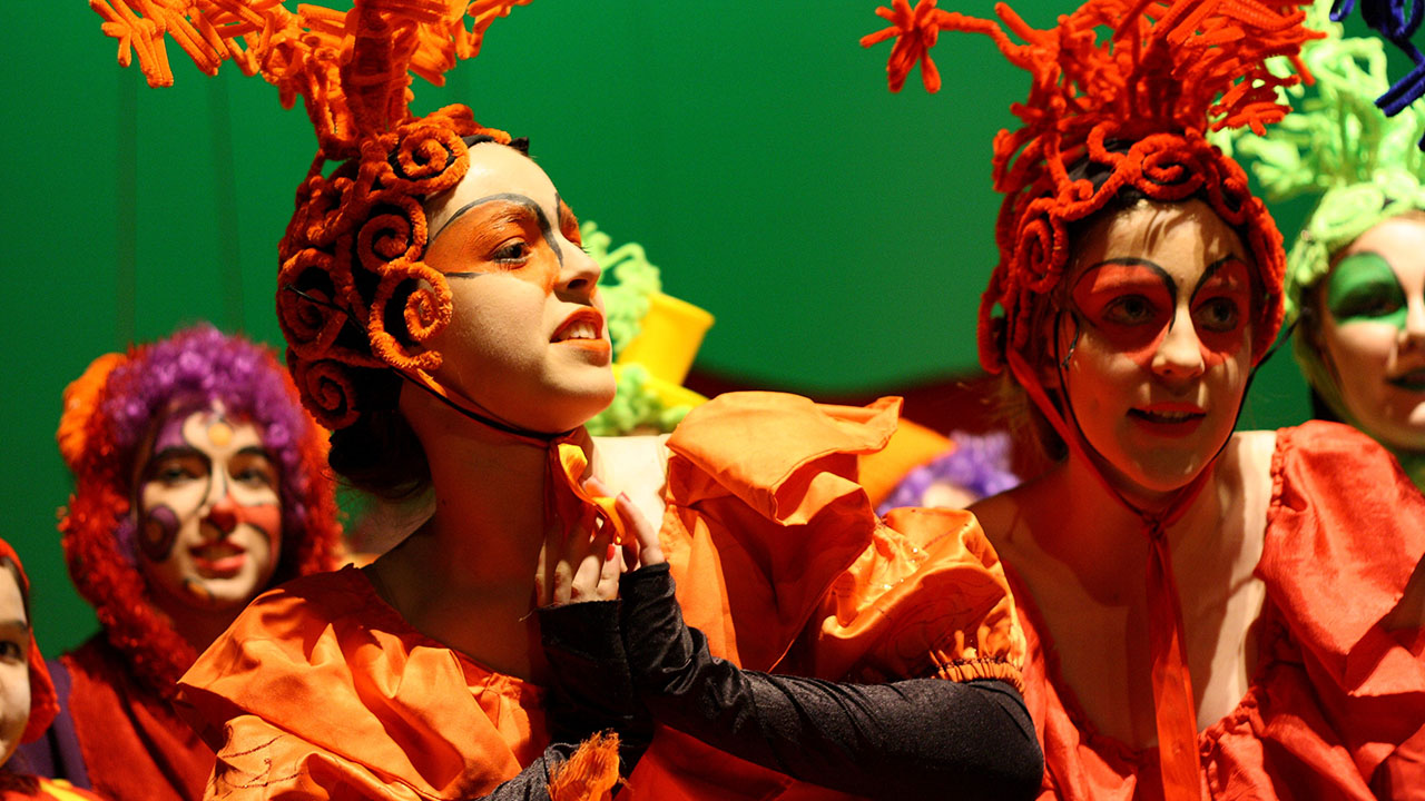 Several students dressed in costumes with orange feathers on their heads and faces painted as if in the circus