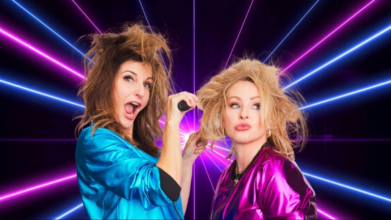 The #IMOMSOHARD duo Kristin Hensley and Jen Smedley doing each others hair poorly in front of an 80s neon laser background