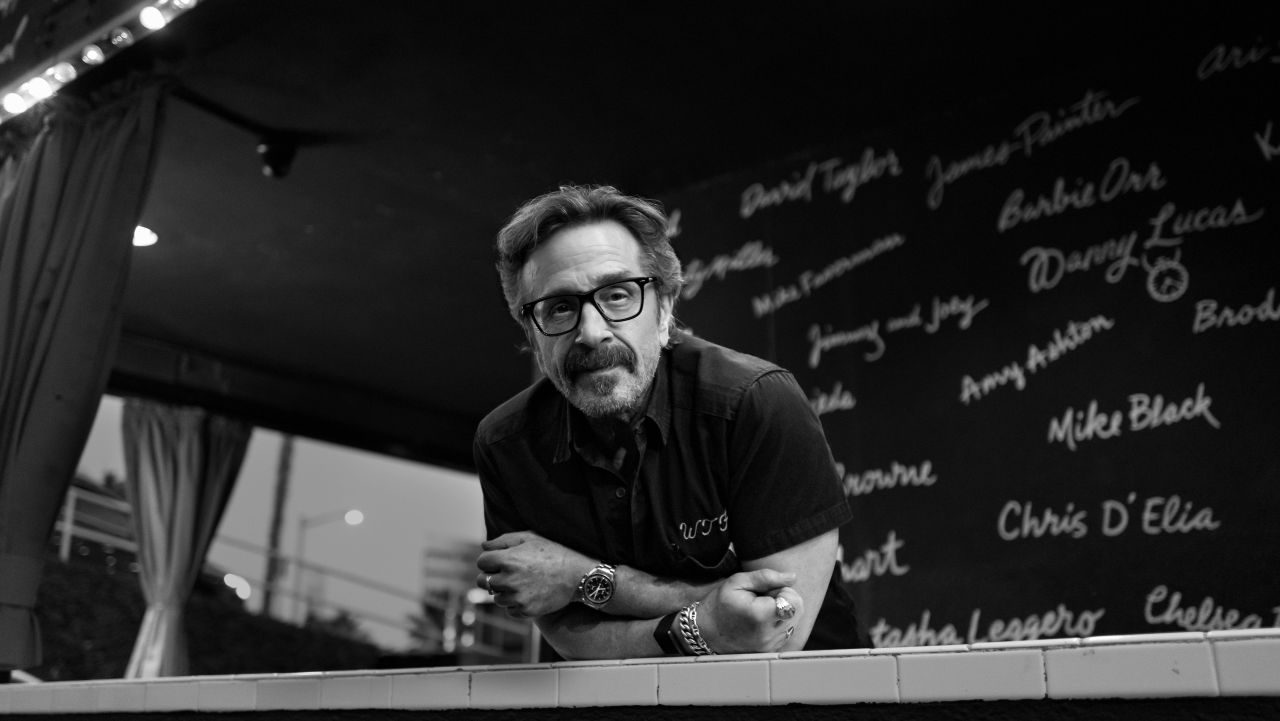 Black and white image of Marc Maron leaning on a white tile ledge at a comedy festival