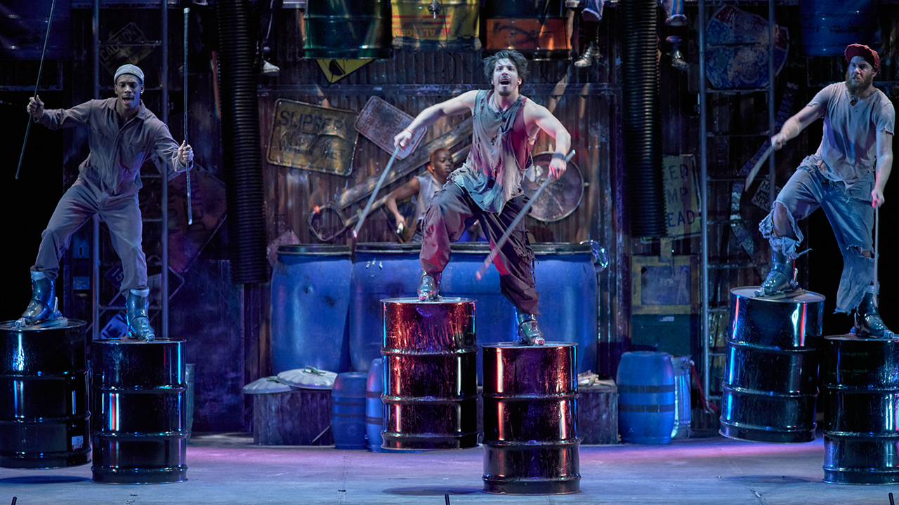 Three members of STOMP pictured on stage with large drum sticks and oil barrels attached to their feet