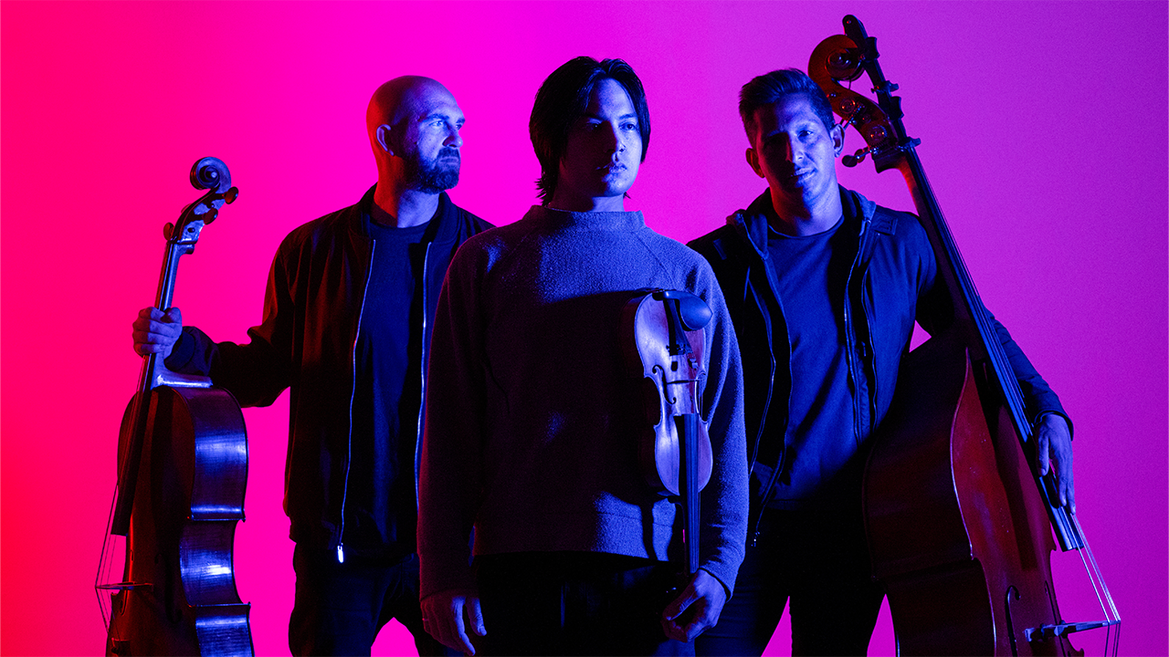 The band Simply Three with their string instruments in front of a dramatic pink/purple background