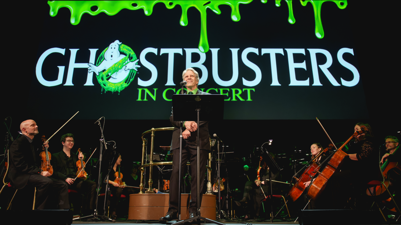 The conductor from Ghostbusters in Concert stands facing the audience with the Ghostbusters logo in the backgrouns