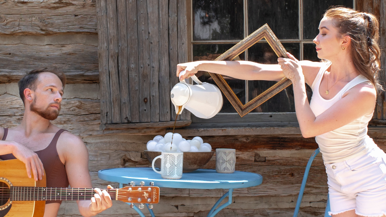 A Dancer pours coffee into a mug and another Dancer observes while holding a guitar.