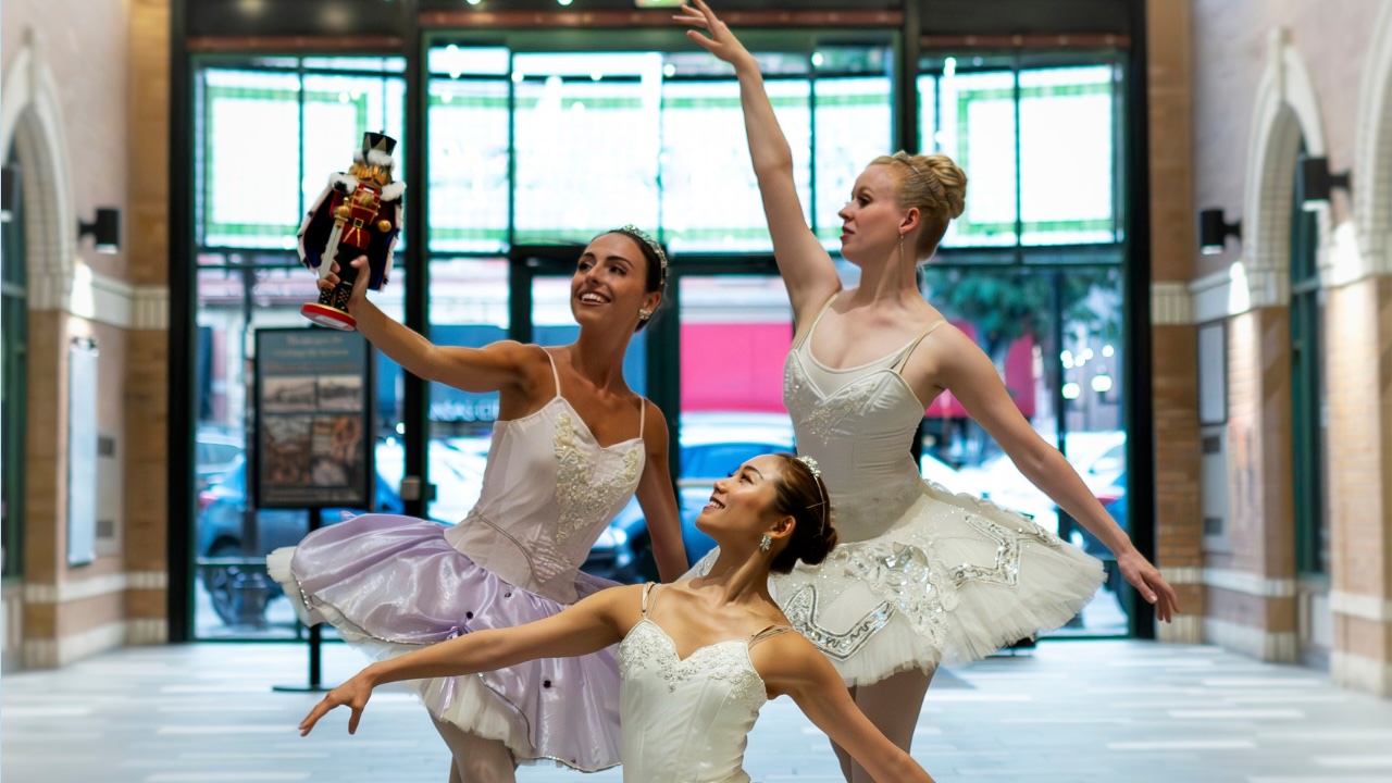 Three ballerinas in white and purple tutus looking at a nutcracker doll in an indoor courtyard.