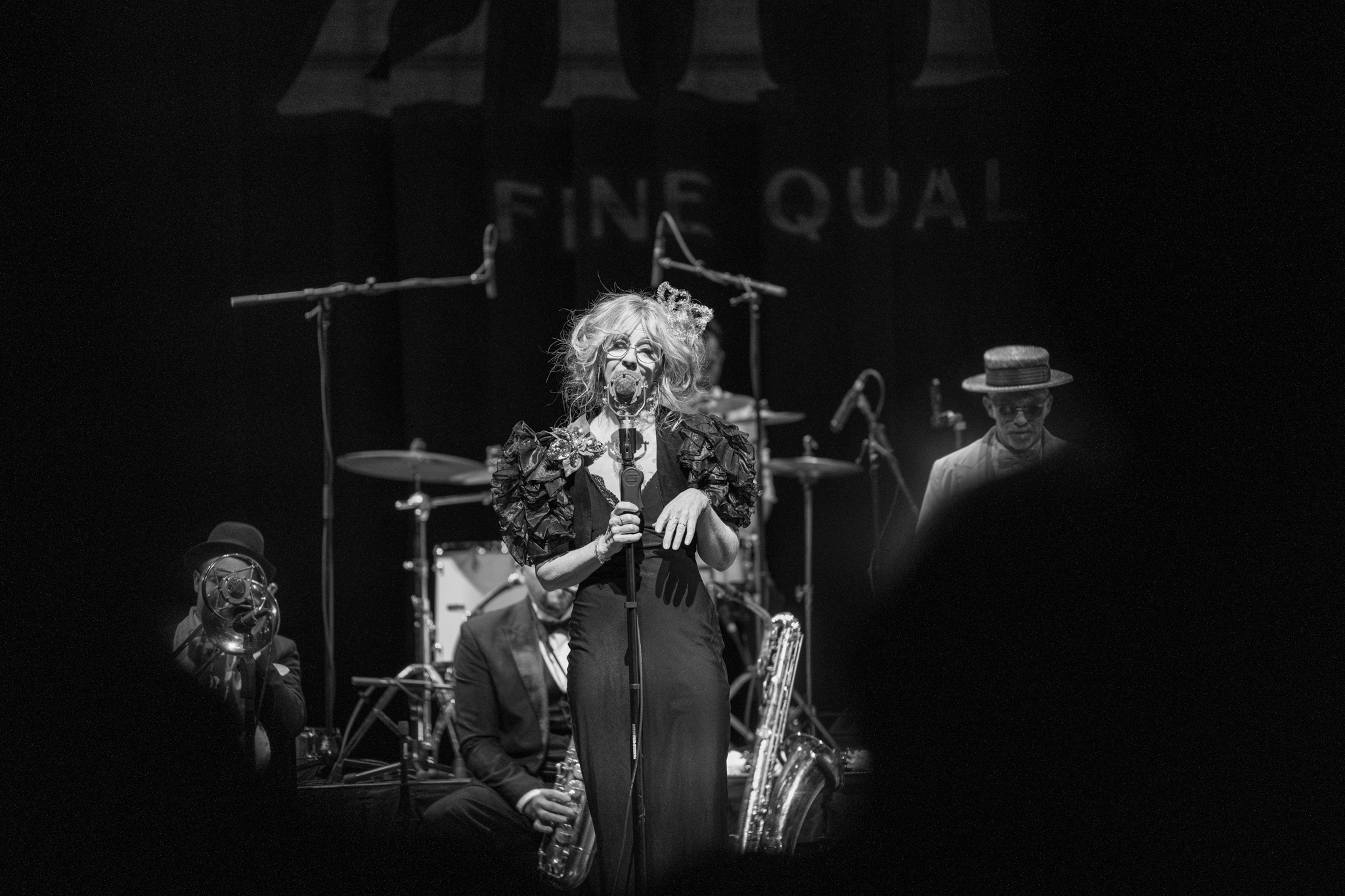 Woman singing into a microphone on stage with backup musicians behind her.