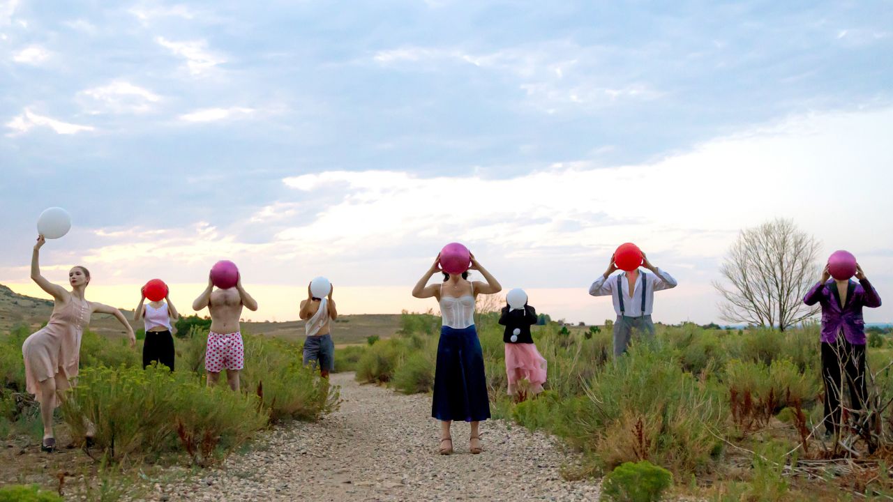 8 Dancers stand in a field with balloons in their mouths, covering their faces. One dancer has the balloon in her hand and makes a pose.
