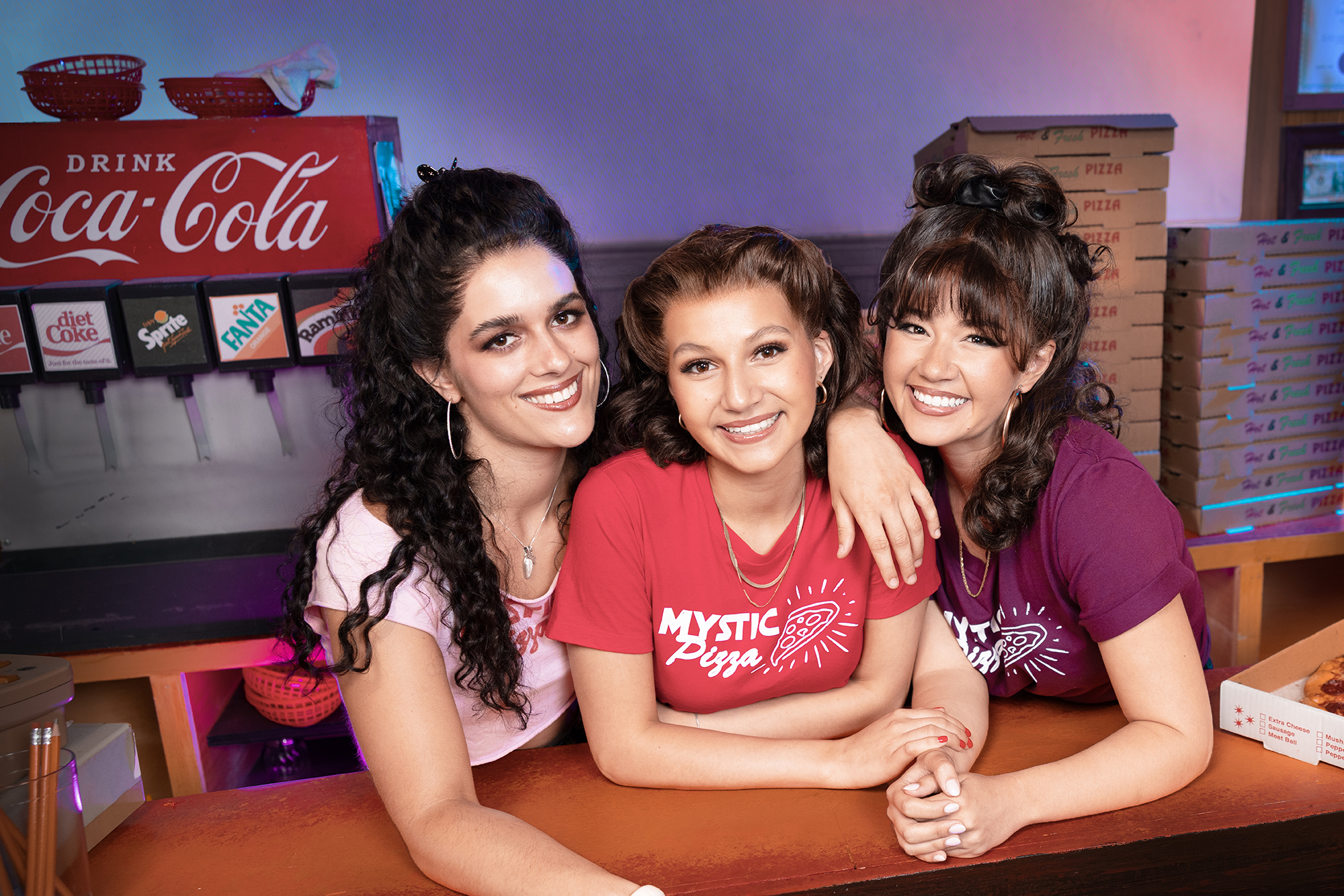 Three actresses in Mystic Pizza shirts pose behind a counter