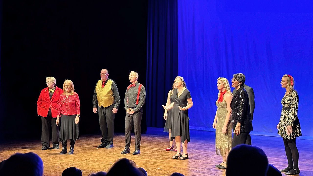 Ten people standing on a stage singing with a blue and black back drop.