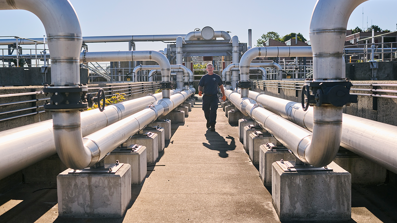 Wastewater utility worker walks on a concrete path at an outdoor facility lined with silver-colored utility pipes on either side.